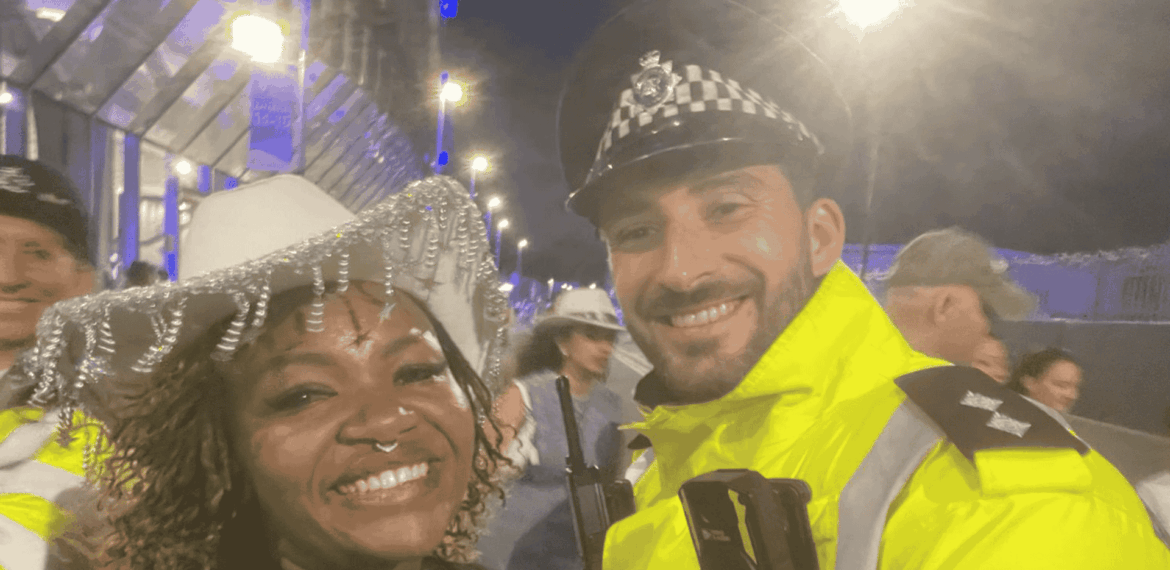 A smiling woman in a white decorative hat, channeling Beyhive energy, stands next to a cheerful police officer in a high-visibility yellow jacket at night, with other people and bright lights in the background.