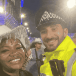 A smiling woman in a white decorative hat, channeling Beyhive energy, stands next to a cheerful police officer in a high-visibility yellow jacket at night, with other people and bright lights in the background.