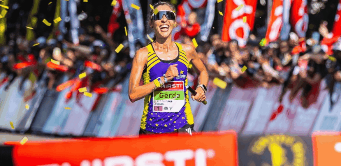 A female runner, Gerda Steyn, smiles as she crosses the finish line, surrounded by cheering crowds and confetti. She wears sunglasses, a purple and yellow athletic outfit, and a race bib reading Gerda.