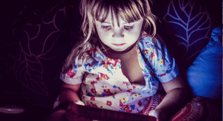 A young child in colorful pajamas sits in dim lighting, illuminated by the glow of a tablet screen, with a patterned cushion and dark leaf-patterned background behind them.