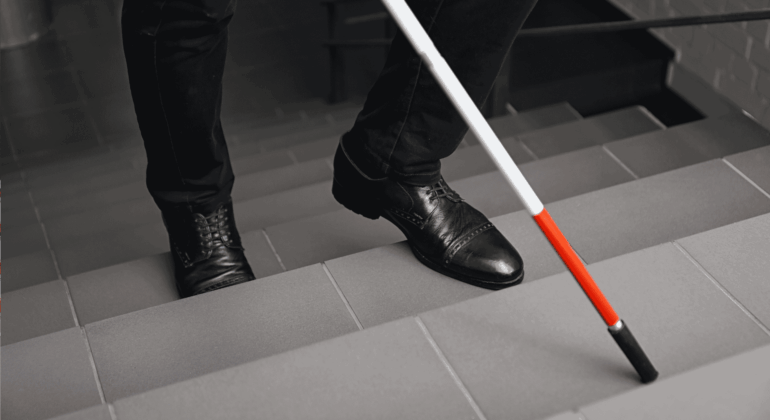A person wearing black shoes and pants uses a white cane with a red tip to navigate down a tiled staircase.