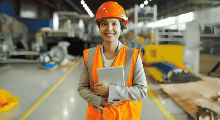 A smiling woman wearing an orange safety vest and hard hat stands in a warehouse, holding a tablet. The background shows industrial equipment and materials, with bright lighting and blurred details.