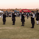 A group of people in kilts and uniforms play bagpipes in a circle on a grassy field during a Highland gathering, with tents and a crowd of onlookers in the background under a clear sky.
