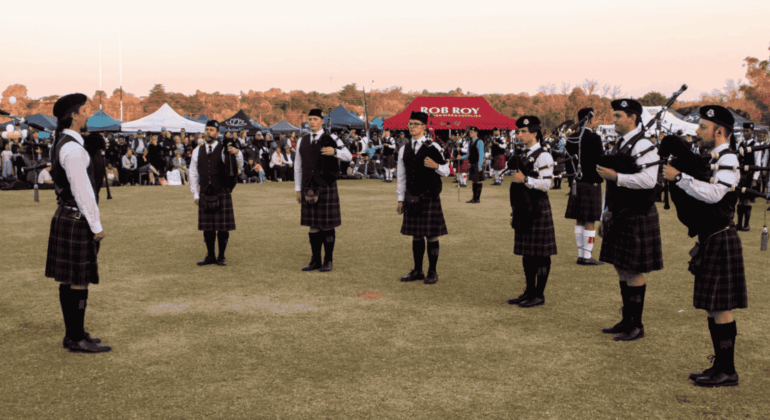 A group of people in kilts and uniforms play bagpipes in a circle on a grassy field during a Highland gathering, with tents and a crowd of onlookers in the background under a clear sky.