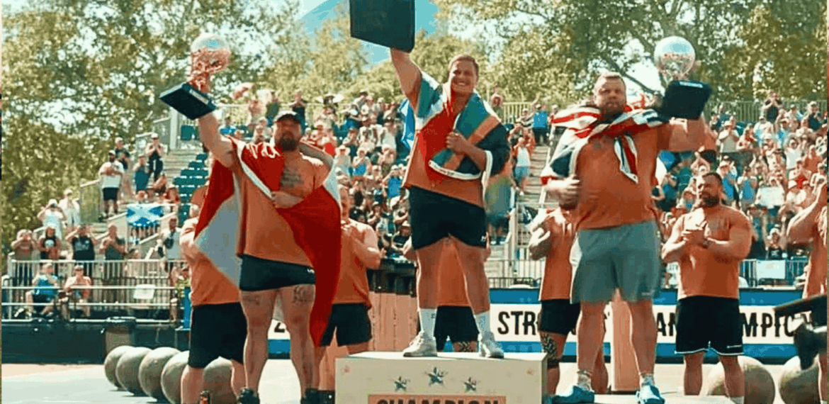 Three men, including Ryno Nel, stand on a winners’ podium at a strongman competition, holding trophies and draped in flags. The central champion raises his trophy high as a cheering crowd and large weights fill the background.