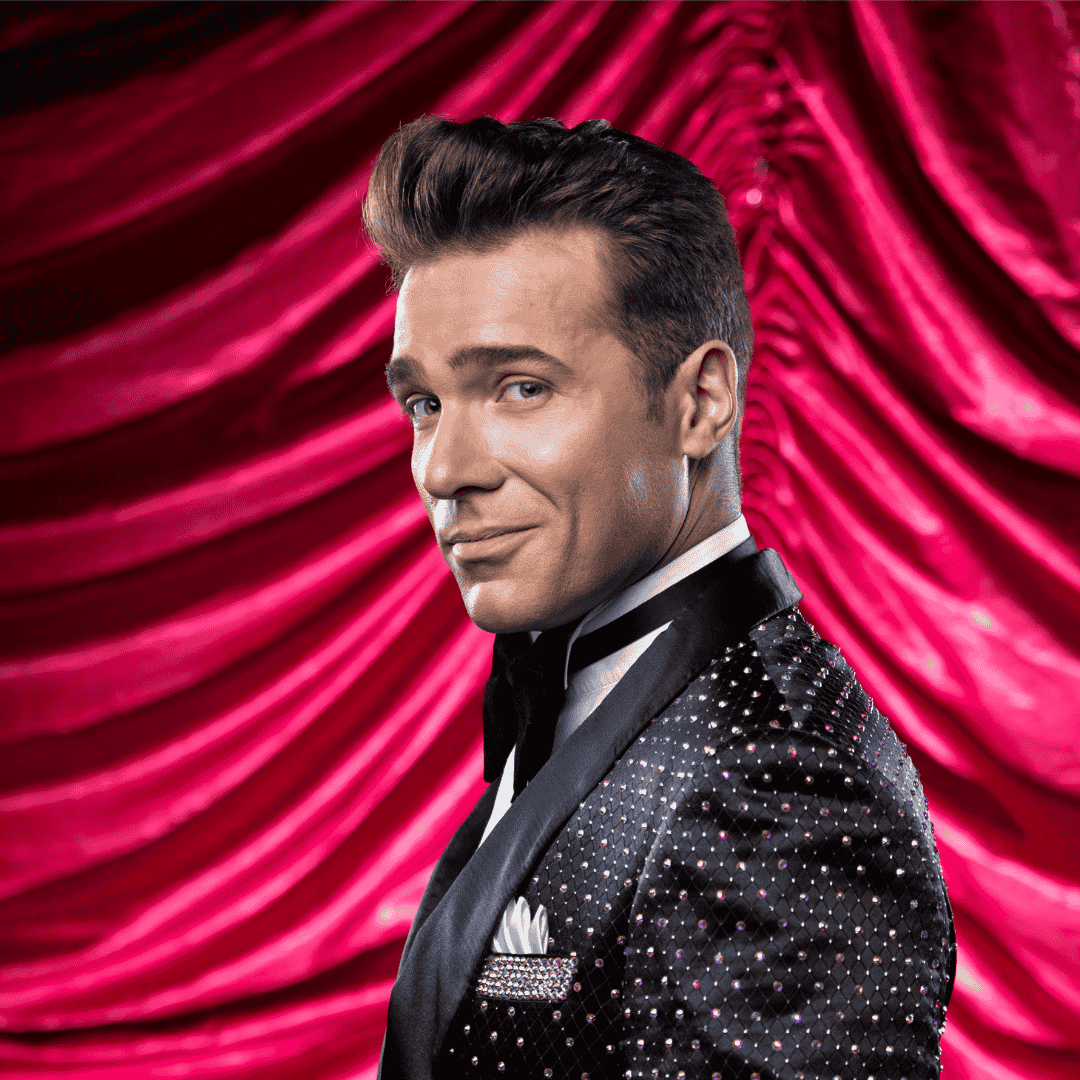 A man in a sparkling black tuxedo with a white shirt and pocket square smiles confidently while standing in front of rich, red, draped velvet curtains.