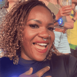 A person with curly hair and a nose ring smiles widely at the camera in their dark top. In the background, others—perhaps fellow Beyhive fans—stand holding drinks as sunlight illuminates this lively outdoor social event.