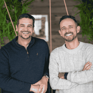 Two men standing side by side, both smiling at the camera. Indoors with green plants hanging in the background, they look like friends ready to expat explore new adventures together—one in a dark sweater, the other in light with arms crossed.