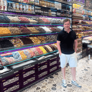 A man in a black shirt and white shorts stands smiling inside a colorful market in Turkiye, surrounded by shelves filled with various spices, dried fruits, and bottles of oil. The floor features a vibrant geometric tile pattern.