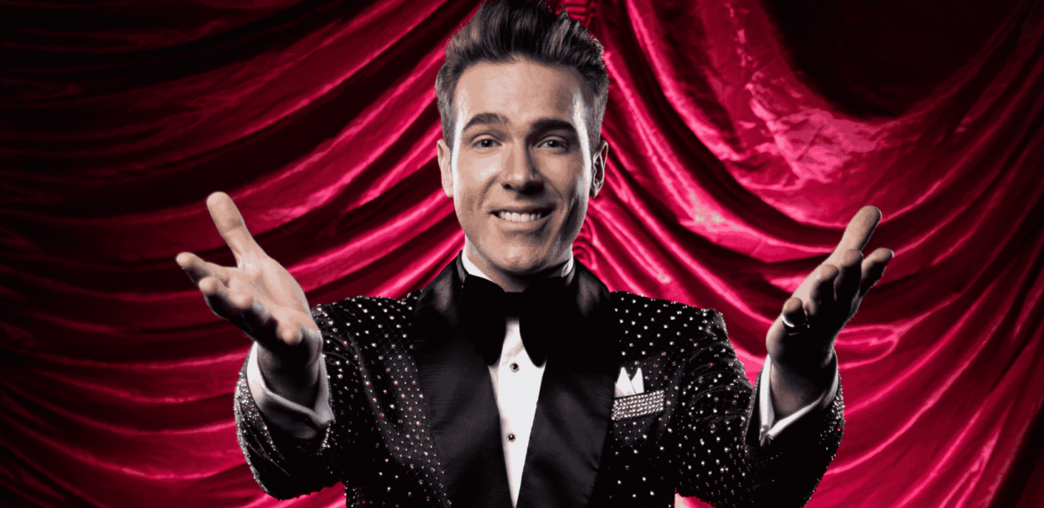 A smiling man in a sparkly black tuxedo with a bow tie stands in front of dramatic red curtains, extending both arms forward in a welcoming gesture, capturing the excitement of a Broadway performance.