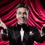 A smiling man in a sparkly black tuxedo with a bow tie stands in front of dramatic red curtains, extending both arms forward in a welcoming gesture, capturing the excitement of a Broadway performance.