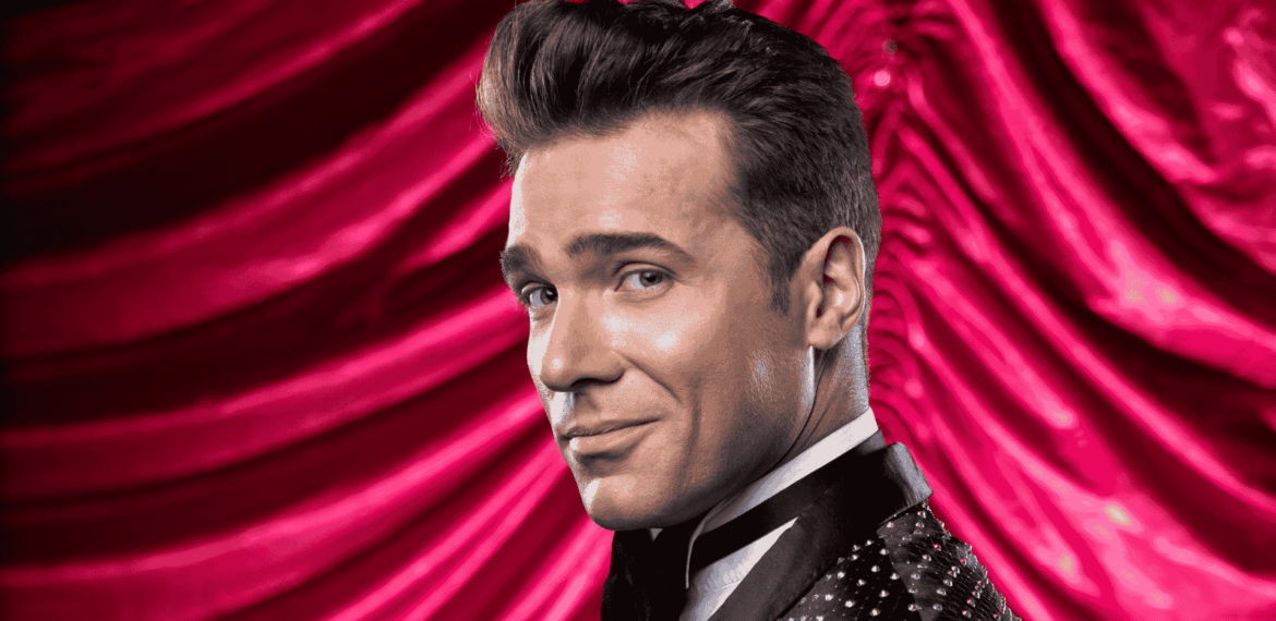 Jonathan Roxmouth, in a sparkling black tuxedo, smiles confidently over his shoulder against a dramatic, red velvet curtain backdrop.