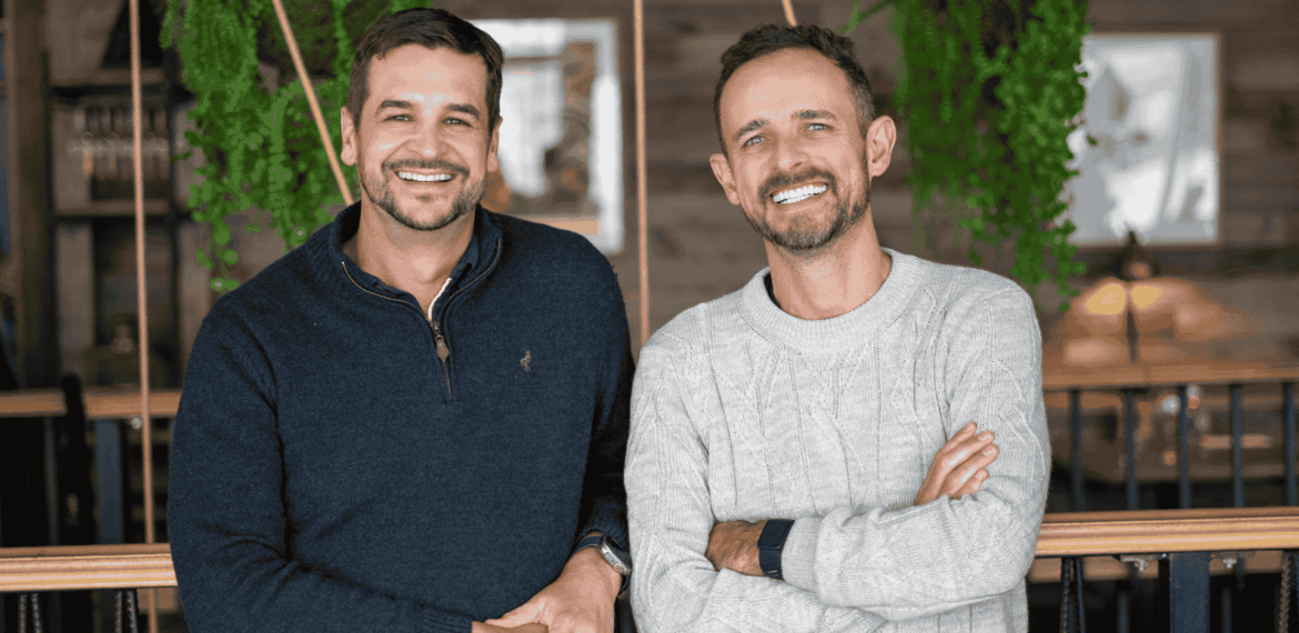 Two men smiling, standing side by side indoors with greenery and wooden decor in the background—perhaps sharing a memorable expat explore experience. One wears a dark sweater, the other a light sweater with arms crossed.