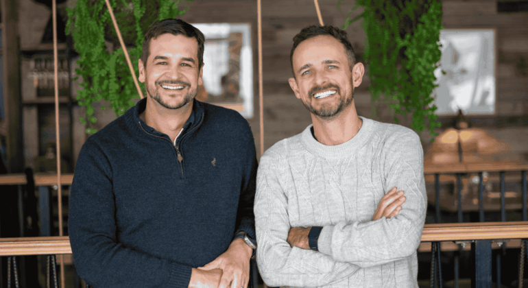 Two men smiling, standing side by side indoors with greenery and wooden decor in the background—perhaps sharing a memorable expat explore experience. One wears a dark sweater, the other a light sweater with arms crossed.
