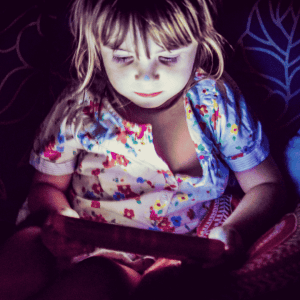 A young child with light hair sits in a dimly lit room, illuminated by the glow of a tablet screen, and wears a colorful floral-patterned shirt while looking intently at the device.