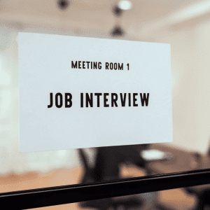 A white paper sign on a glass door reads “Meeting Room 1 Job Interview” in bold black letters, with a blurred office setting visible in the background.