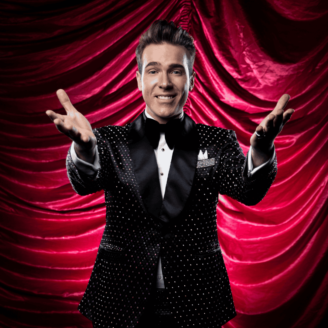 A smiling man in a black tuxedo with white polka dots stands in front of red, draped curtains, holding his arms out as if welcoming or inviting an audience.