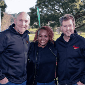 Three people stand close together outdoors, smiling at the camera on their Sunshine tour. They all wear black jackets, with green trees and grass in the background under a clear sky.
