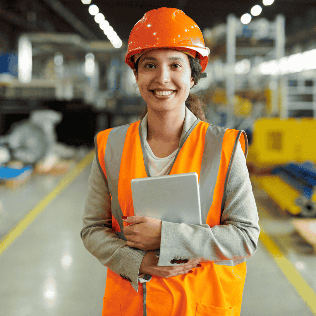 A smiling woman wearing an orange safety helmet and reflective vest holds a tablet while standing in a brightly lit industrial warehouse.