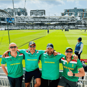 Four smiling men in green Proteas cricket shirts stand arm-in-arm at a cricket stadium, with the field and stands visible behind them under a partly cloudy sky.