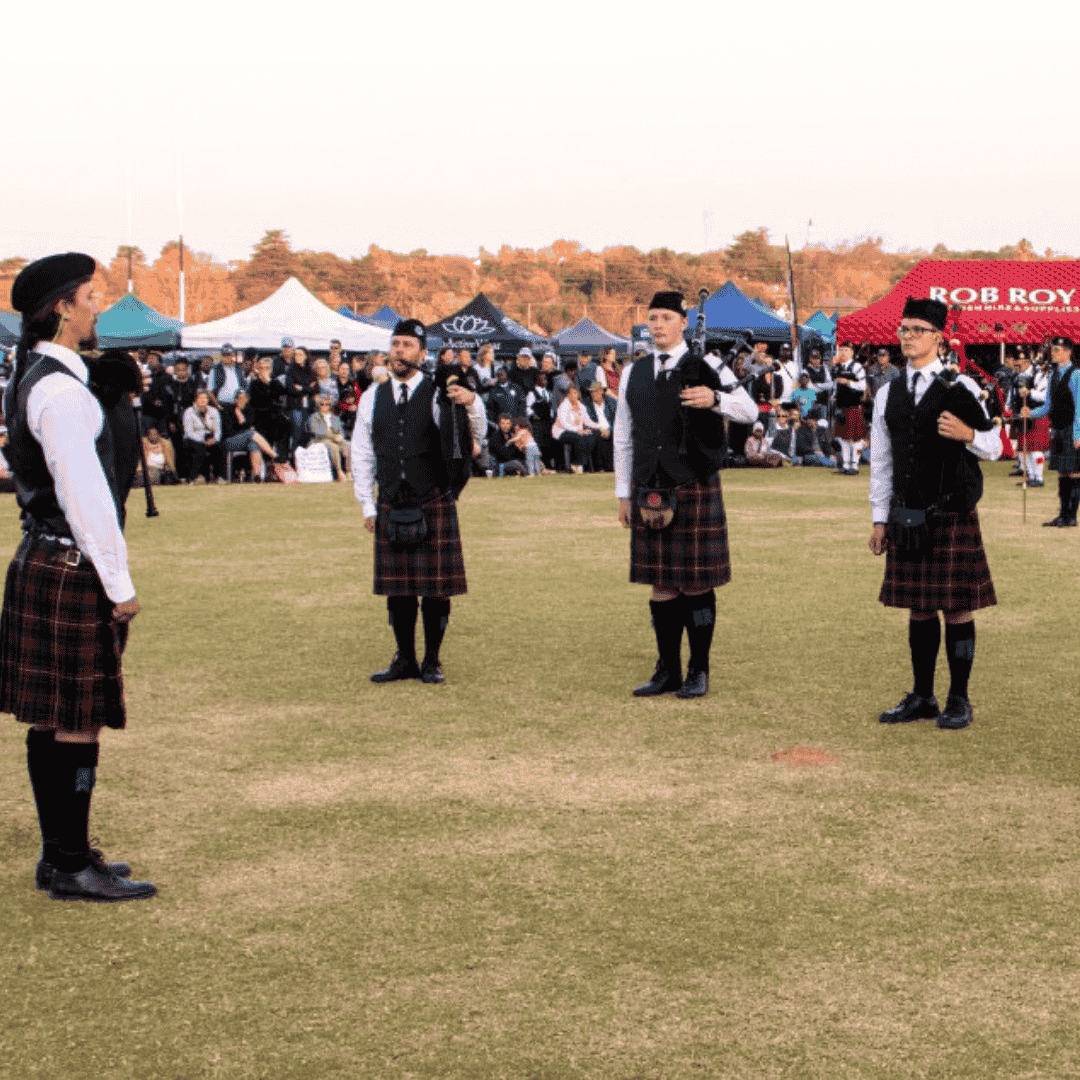Four people wearing traditional Scottish kilts and vests stand in a circle on a grassy field, performing or preparing to perform, with a crowd and colorful tents in the background.