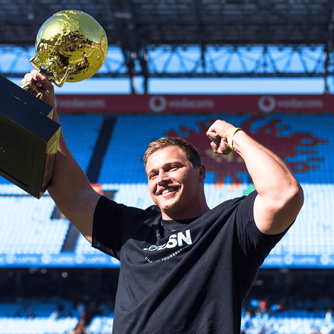 A smiling man in a black T-shirt, identified as Ryno Nel, flexes his arm and lifts a large gold trophy in a stadium with blue seats and a partially filled crowd in the background.
