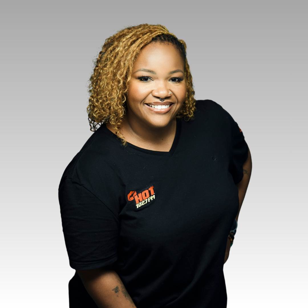 A smiling woman with curly blonde hair, Bunny Majaja, wears a black Hot 102.7 FM t-shirt and stands against a light gray gradient background. She is facing forward with one hand on her hip.