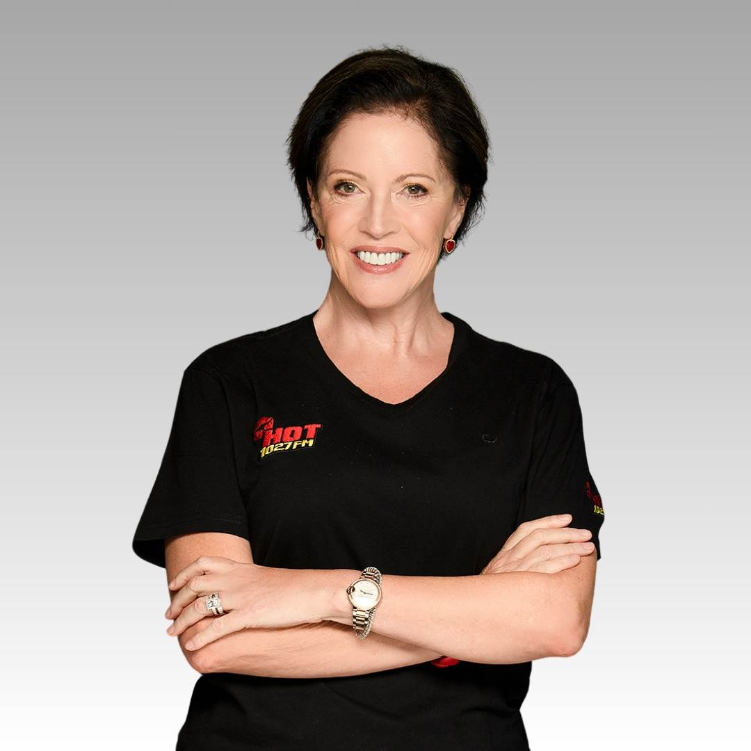 A woman with short dark hair, wearing a black t-shirt with red logo text, smiles with arms crossed against a gray gradient background. She is wearing a watch, rings, and earrings.