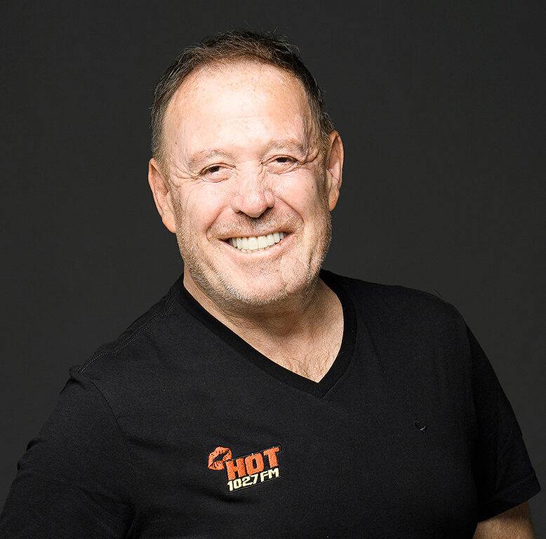 A smiling man in a black t-shirt with the HOT 102.7 FM logo stands against a plain dark background, ready to broadcast live from a special memorial service.