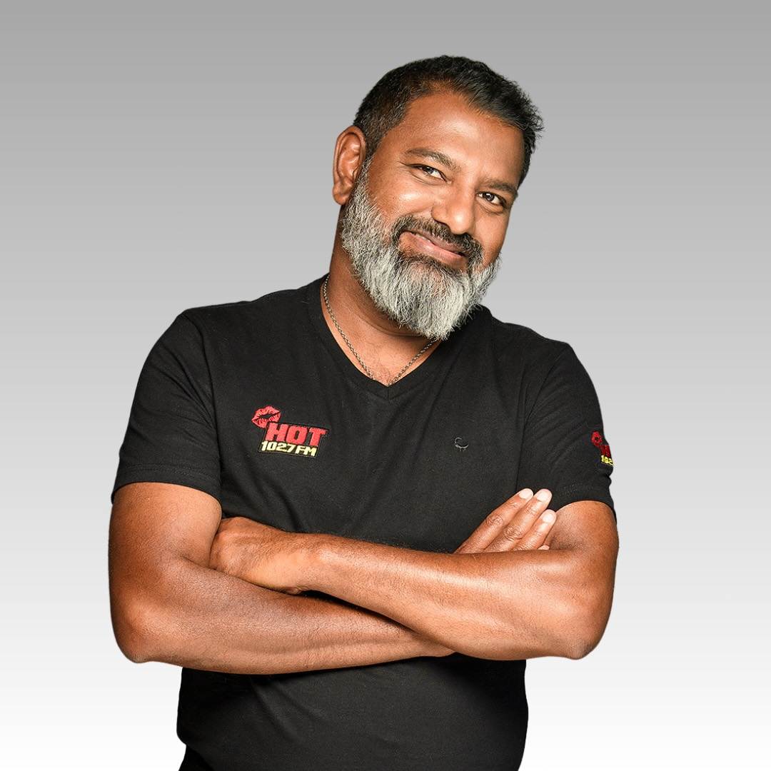 A man with short hair and a salt-and-pepper beard smiles with his arms crossed, wearing a black t-shirt with HOT 102.7 FM logos on the chest and sleeve, standing against a gray gradient background.