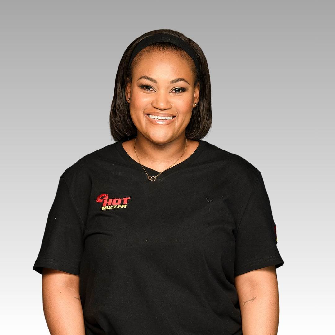 A smiling woman with straight shoulder-length hair wears a black t-shirt with HOT text on it, standing against a plain gray background.