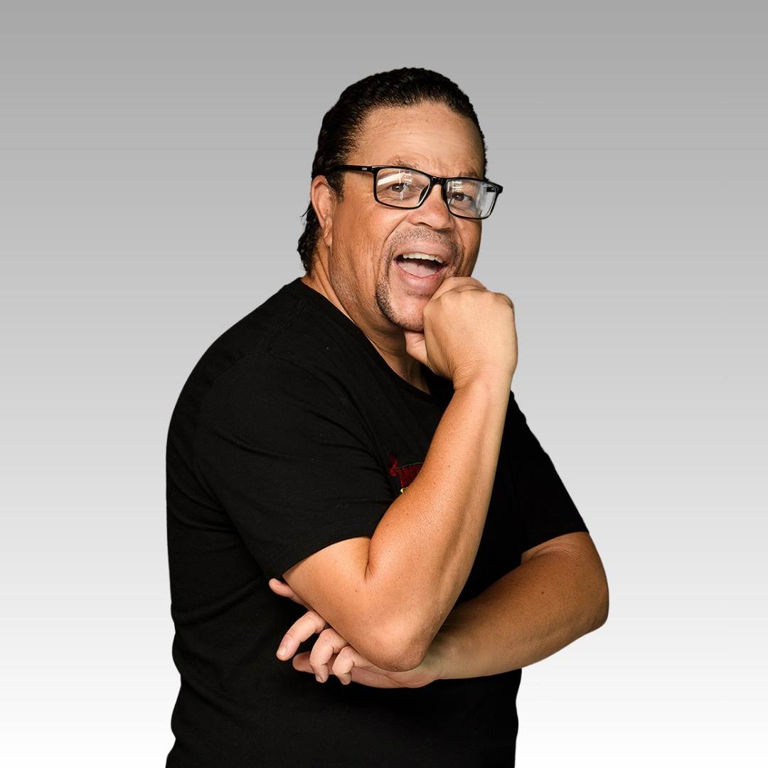 A man with glasses and a black t-shirt poses against a gray gradient background, smiling with his hand resting on his chin and his other arm crossed over his body.