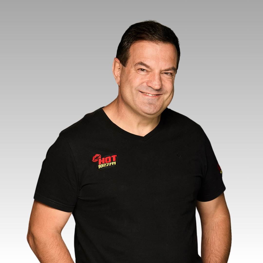 A man with short dark hair smiles while standing against a gray gradient background. He is wearing a black t-shirt with the HOT 102.7 FM logo on the left side of his chest.