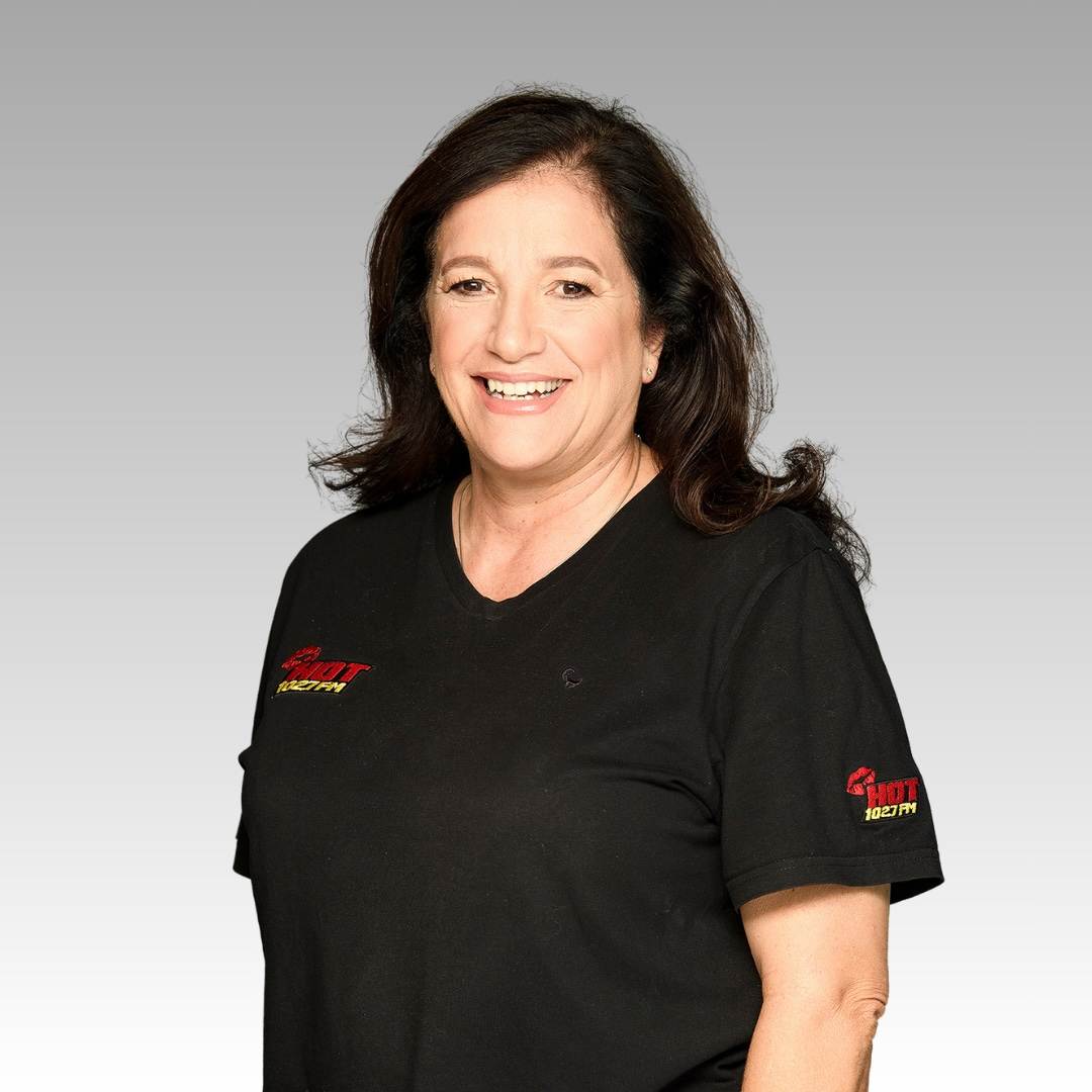 A woman with long dark hair smiles at the camera. She is wearing a black T-shirt with red and yellow logos on the chest and sleeve, standing in front of a plain, light gray background.