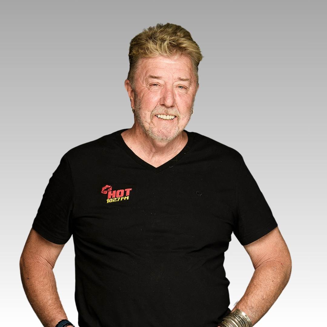 A smiling older man with light hair and a beard wears a black HOT 102.7FM T-shirt, a watch, and bracelets, standing against a plain gray gradient background.