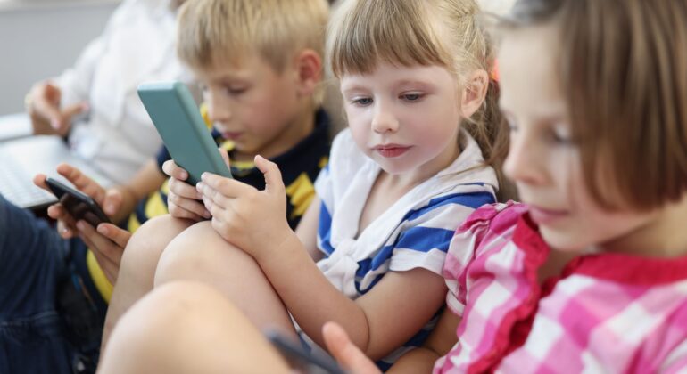 Three young children sit closely together in a bright indoor setting, each focused on a smartphone or tablet screen. Dressed in casual striped clothing, they appear fully engaged with their devices, absorbed in their screen time.