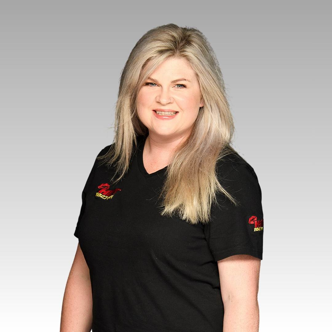 A smiling woman with long blonde hair wearing a black T-shirt with red and yellow logos on the chest and sleeve stands against a light gray gradient background.
