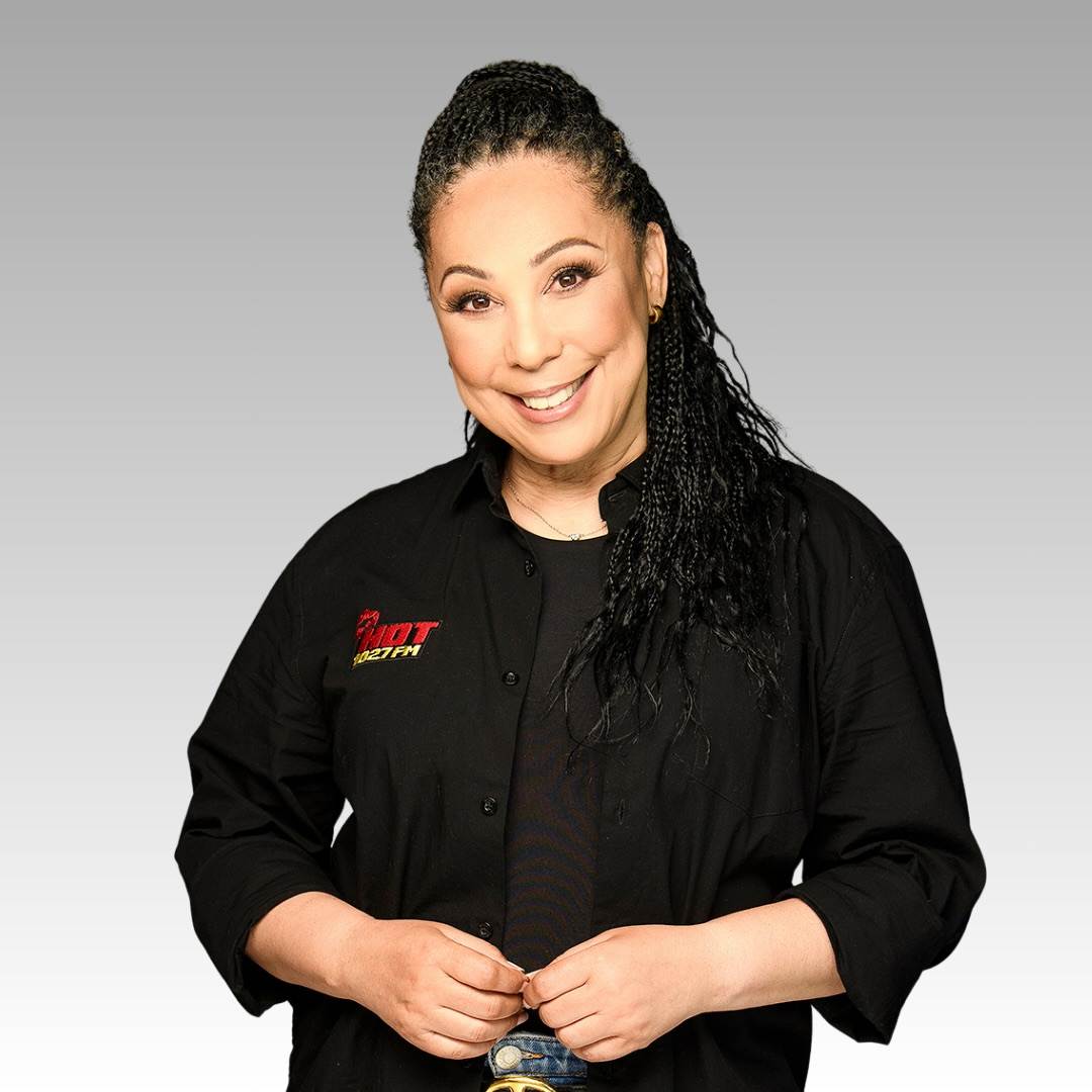 A smiling woman with braided hair wearing a black button-up shirt with red and yellow embroidery stands against a gray gradient background.
