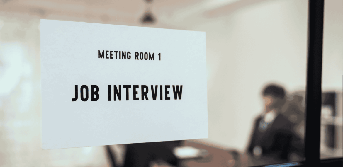 A sign reading Meeting Room 1 Job Interview is posted on a glass door. In the blurry background, two people are seated at a table, suggesting an interview is in progress.