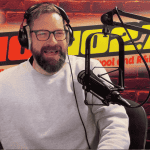 A bearded man wearing glasses and a light gray sweater speaks into a microphone in a radio studio with a red brick wall, HOT 96.7 FM logo, and a red lips graphic in the background. Ian F