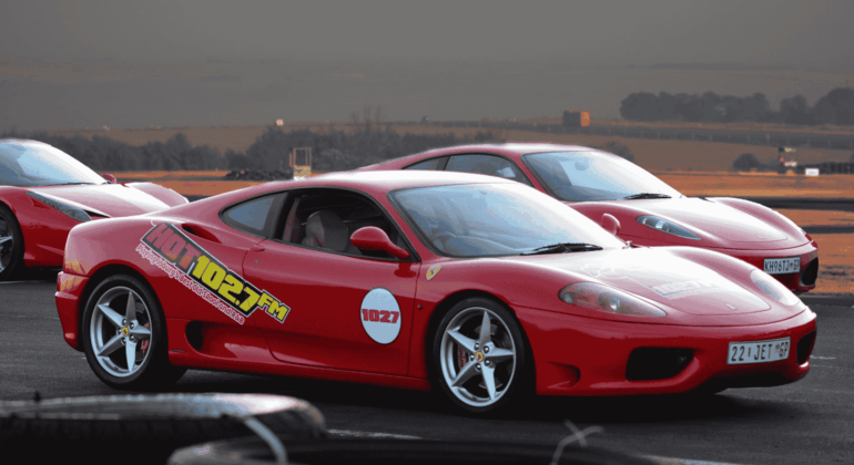 A red Ferrari sports car with 102.7 FM decals parked on a racetrack, with another red Ferrari and open landscape in the background under a cloudy sky.