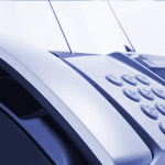 Close-up of a fax machine with paper inserted, showing its keypad and control panel. The image has a blue tint and focuses on the device’s buttons and curved edges.
