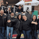 A group of people wearing black jackets stand and smile in front of an outdoor radio station setup, with some people sitting or standing behind a DJ booth under a white umbrella. Everyone looks happy and cheerful.