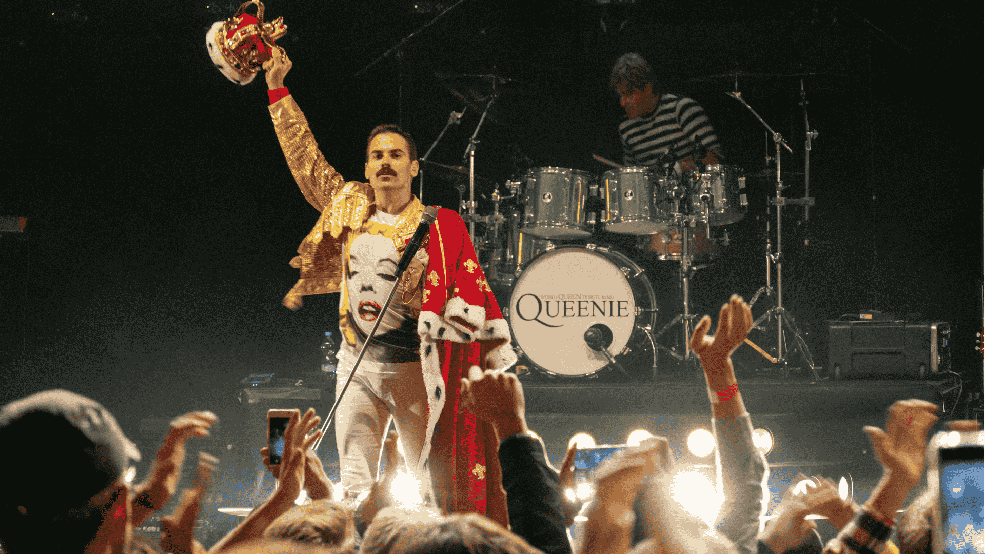 A performer dressed as Freddie Mercury holds a crown aloft on stage, wearing a gold jacket and crown robe. A drummer plays behind him, and the crowd raises their hands. The drum set proudly displays the word QUEENIE.