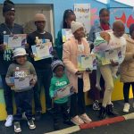 A group of children and teens stand outdoors, holding certificates and smiling for a photo. They are dressed warmly in jackets and hats, with colorful walls behind them. An adult’s hand is visible, taking the picture.