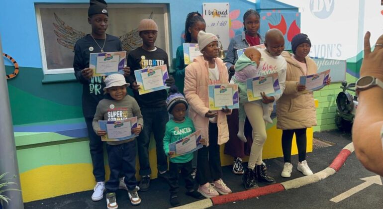 A group of children and teens stand outdoors, holding certificates and smiling for a photo. They are dressed warmly in jackets and hats, with colorful walls behind them. An adult’s hand is visible, taking the picture.