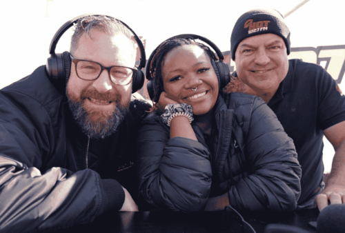 Three radio hosts wearing headphones smile at the camera while sitting close together at a broadcasting table, with a station logo visible in the background.
