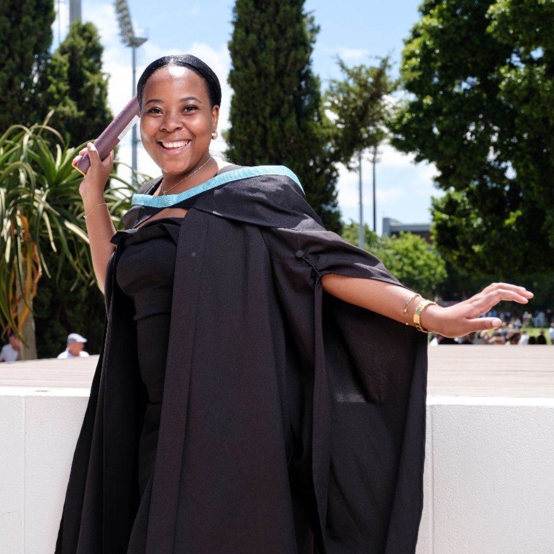 A smiling woman in a graduation gown and cap holds a diploma and poses outdoors on a sunny day, with trees and a sports field in the background.