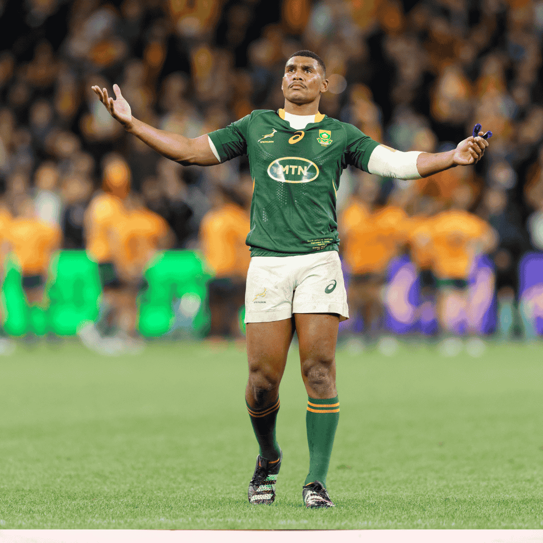 A rugby player in a green Springboks jersey stands on a field with arms outstretched, looking upwards. Players in orange uniforms are blurred in the background with a crowd watching.