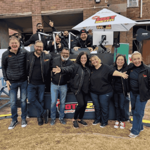 A group of nine smiling people wearing black jackets pose together in front of a radio station outdoor booth with HOT 91.9 FM branding, in an urban setting.